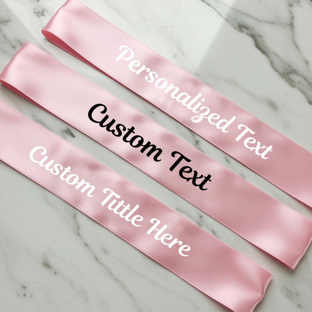 Pink satin sashes with customizable text on a marble surface
