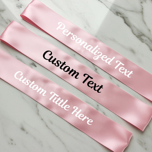 Pink satin sashes with customizable text on a marble surface
