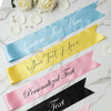 Four customizable satin ribbons in blue, yellow, pink, and black on a marble surface with white flowers.